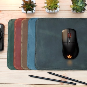 May include: A dark green leather mouse pad with a black mouse and the initials "AM" embossed in the bottom right corner. The mouse pad is surrounded by six smaller leather mouse pads in different colors, including dark green, red, brown, and blue.