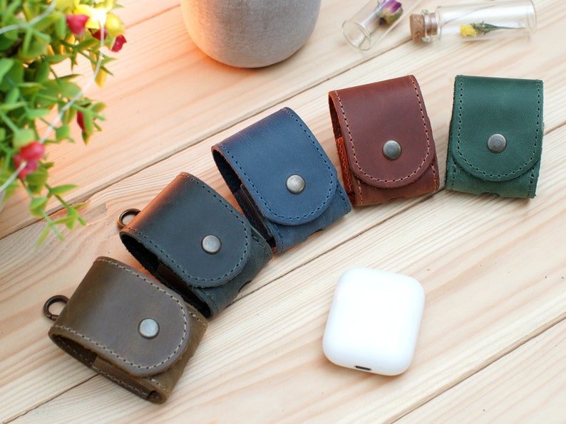 May include: Five leather cases for wireless earbuds in different colors: brown, dark blue, light blue, green, and dark brown. The cases have a snap closure and a small metal loop.