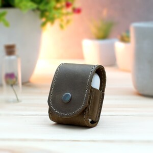 May include: A brown leather case for wireless earbuds. The case has a flap that closes with a snap and a small metal button on the front.