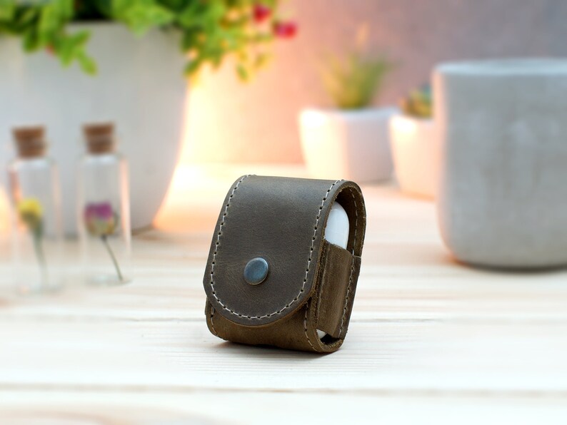 May include: A brown leather case for wireless earbuds. The case has a flap that closes with a snap and a small metal button on the front.