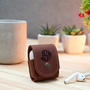 May include: A brown leather case for wireless earbuds with a monogrammed initial "JSM" in black. The case has a button closure and is sitting on a wooden surface.