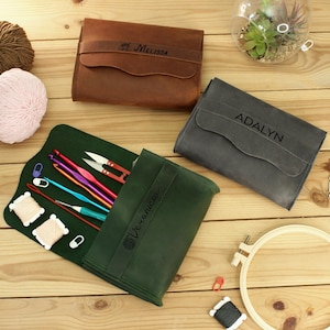 May include: Three leather needlepoint cases with different names on them. The green case is open and has knitting needles and other tools inside. The case is on a wooden surface.