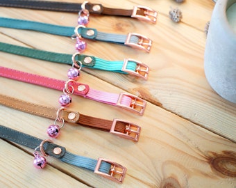Könnte beinhalten: Ein Set aus acht bunten Katzenhalsbändern mit rosa Glöckchen. Die Halsbänder sind aus verschiedenen Materialien gefertigt, darunter Leder, Wildleder und Stoff. Die Halsbänder sind in einer Reihe auf einer Holzoberfläche angeordnet.