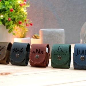 May include: Five personalized leather Airpod cases in different colors. The cases are brown, dark green, dark blue, and light blue. The cases are engraved with the names "SAR", "Rich", "ANR", "Robert", and "James".