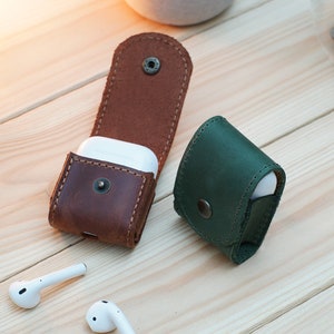 May include: Two leather cases for wireless earbuds. One case is brown leather and the other is green leather.