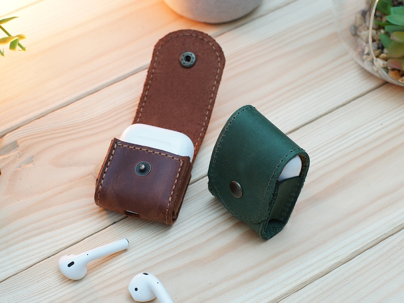 May include: Two leather cases for wireless earbuds. One case is brown leather and the other is green leather.