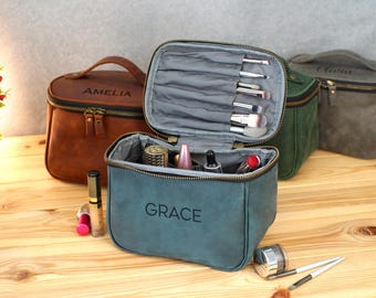May include: A collection of personalized makeup bags in various colors, including brown, green, and teal. The teal bag is open, displaying makeup brushes and products. Each bag has a name printed on it.