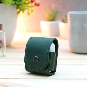 May include: A dark green leather case for wireless earbuds. The case has a stitched seam around the edge and a metal button on the front.