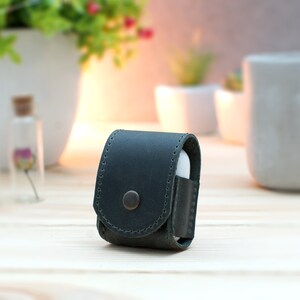 May include: A dark green leather case for wireless earbuds. The case has a snap closure and a small button on the front.