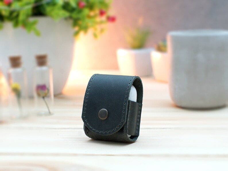 May include: A dark green leather case for wireless earbuds. The case has a snap closure and a small button on the front.