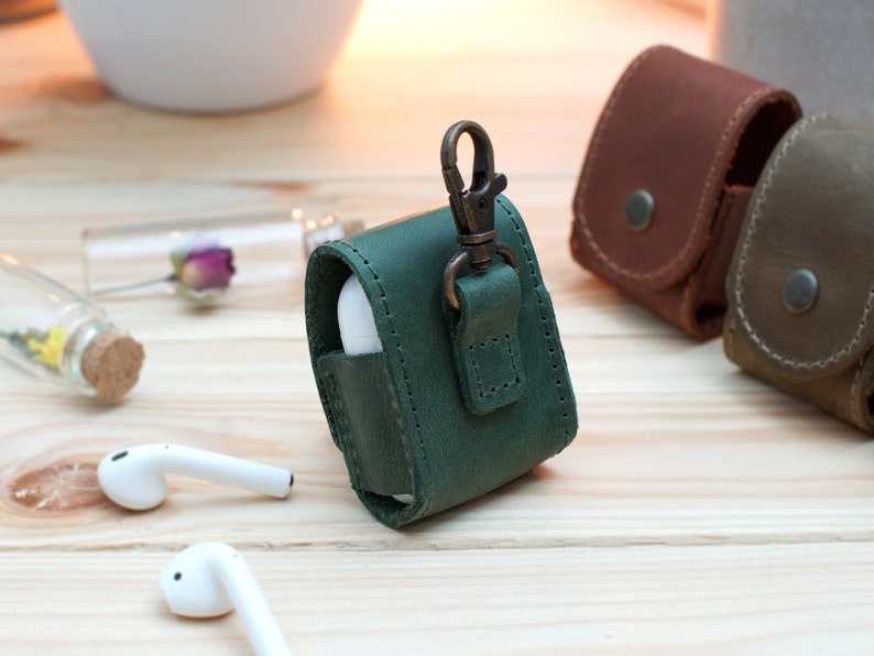 May include: Green leather case with a metal clasp for AirPods. The case has a strap for attaching to a keychain.