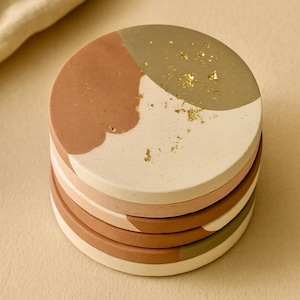 May include: A stack of six round coasters. Each coaster is divided into three sections of color: terracotta, off-white, and olive green. Gold flecks are scattered across the top coaster. The coasters are made of a solid material.