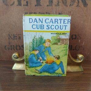 May include: Vintage book cover featuring a colorful illustration of three boys in blue scout uniforms gathered around a campfire in a forest setting. The title reads "Dan Carter Cub Scout" by Mildred A. Wirt.