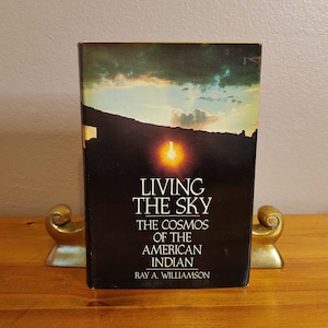 May include: A book titled "Living the Sky: The Cosmos of the American Indian" by Ray A. Williamson. The cover features a sunset over a mountain range with a bright orange sun.