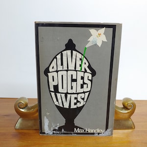 May include: A gray book cover with black and white text that reads "Oliver Poques Lives!"  The text is in a stylized font and is surrounded by a black silhouette of a vase with a white flower on top. The author's name, "Max Handley", is printed in black at the bottom of the cover.