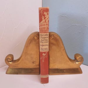 May include: A vintage book titled "Five Little Peppers And How They Grew" stands between two gold-colored bookends. The book has a red cover with white illustrations and text. The bookends have a curved design.