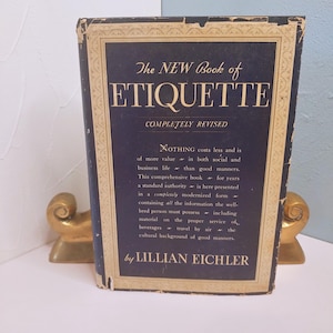 May include: A vintage book titled "The NEW Book of ETIQUETTE" with a navy blue cover and gold lettering. The book is propped up by two gold-colored bookends. The book's cover includes text about social and business etiquette.