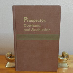 May include: A brown book with the title "Prospector, Cowhand, and Sodbuster" in gold lettering. The book has a green and brown striped design on the cover.