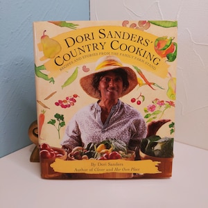 May include: A cookbook titled "Dori Sanders' Country Cooking" with a woman wearing a straw hat on the cover. The cover features illustrations of vegetables and fruits. The book is by Dori Sanders, author of "Clover and Her Own Place."