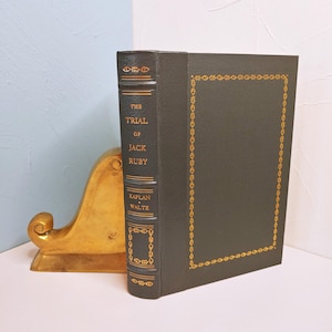 May include: A dark green hardcover book titled The Trial of Jack Ruby by Kaplan and Waltz, featuring gold foil lettering and decorative border detailing on the spine and front cover, positioned next to a curved gold-toned metal bookend.