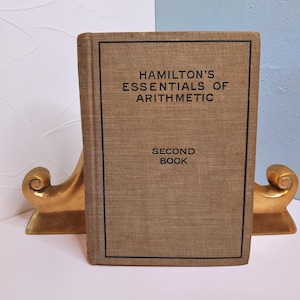 May include: A vintage book titled "Hamilton's Essentials of Arithmetic, Second Book" stands upright between two gold-colored bookends. The book has a tan cover with black lettering and a black border. The bookends have a decorative, curved design.