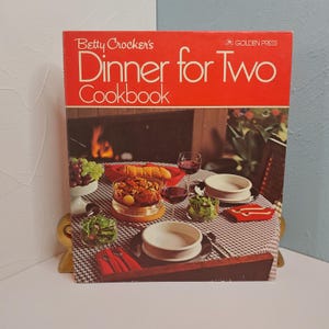 May include: A vintage cookbook titled "Betty Crocker's Dinner for Two Cookbook" with a red cover. The cover features the title in white and a photograph of a dinner table set for two, with food and wine.