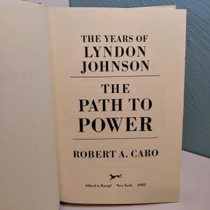 May include: An open book with a cream-colored page featuring the title "THE YEARS OF LYNDON JOHNSON THE PATH TO POWER" in bold black font. The author's name, Robert A. Caro, is below the title. The book was published in 1982 by Alfred A. Knopf, New York.