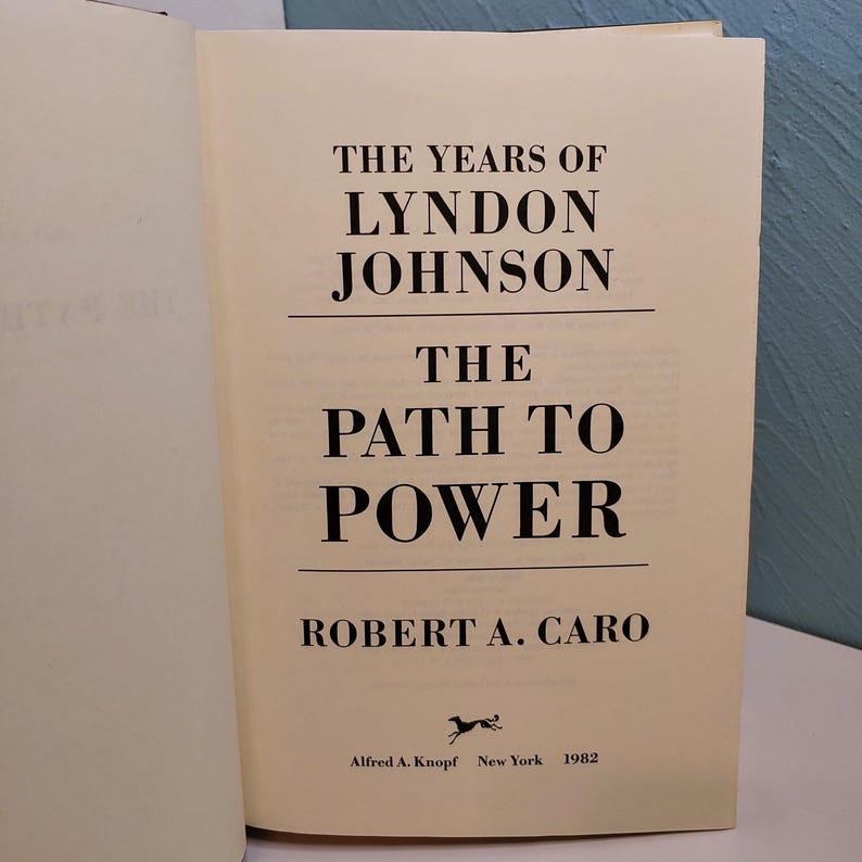 May include: An open book with a cream-colored page featuring the title "THE YEARS OF LYNDON JOHNSON THE PATH TO POWER" in bold black font. The author's name, Robert A. Caro, is below the title. The book was published in 1982 by Alfred A. Knopf, New York.