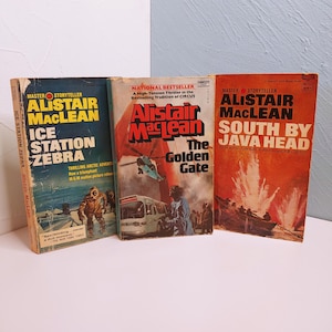 May include: Three vintage paperback books by Alistair MacLean. The book titles are "Ice Station Zebra", "The Golden Gate", and "South by Java Head". The covers feature illustrations and text in various colors, including blue, orange, and white. The books are displayed on a white surface.