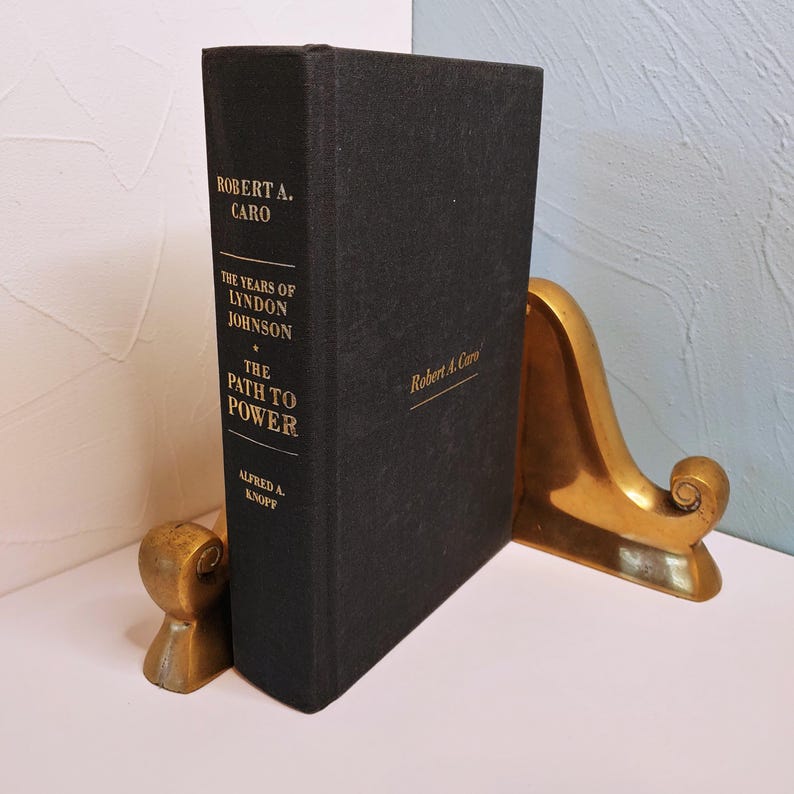 May include: A hardcover book titled "The Years of Lyndon Johnson: The Path to Power" by Robert A. Caro, held between two brass bookends. The book has gold lettering on a black cover. The bookends have a decorative, curved design.