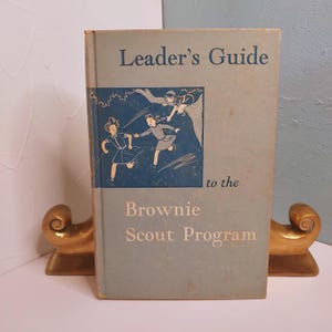 May include: A vintage book titled "Leader's Guide to the Brownie Scout Program." The cover is light blue with illustrations of girls and text in white and blue. The book is propped up by gold-colored bookends.