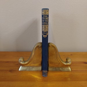 May include: A blue hardcover book titled "The Story of the Bible" with gold lettering and decorative borders. The book is standing upright in a pair of gold metal bookends.