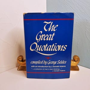 May include: A hardcover book titled "The Great Quotations" with a blue cover and white lettering. The book is compiled by George Seldes and includes an introduction by J. Donald Adams. The book is flanked by two gold-colored bookends.