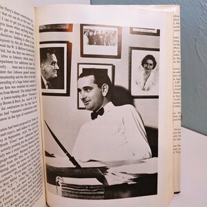 May include: Black and white photograph of a man in a white shirt and bow tie, seated at a desk with a pen. Several framed portraits hang on the wall behind him. The image is from a book.