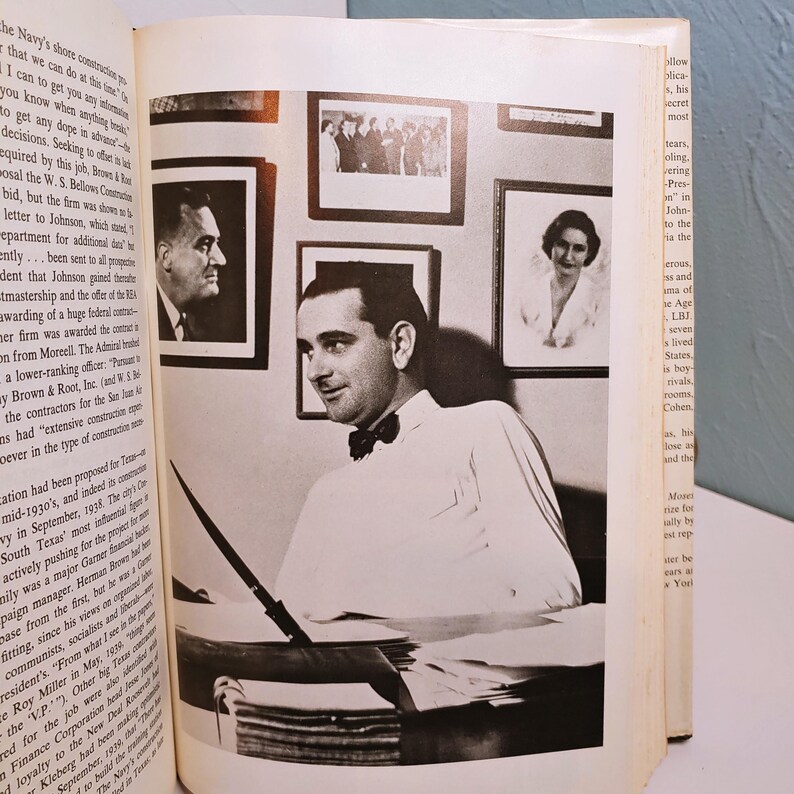 May include: Black and white photograph of a man in a white shirt and bow tie, seated at a desk with a pen. Several framed portraits hang on the wall behind him. The image is from a book.