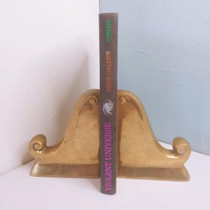 May include: A pair of gold-colored bookends with a decorative, curved design. A book titled "Violent Universe" by Nigel Calder, published by Viking, stands between the bookends. The book has a brown cover with purple and green text.