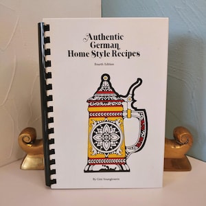 May include: A white cookbook titled "Authentic German Home Style Recipes" with a black spiral binding. The cover features a colorful illustration of a German beer stein. The book is propped up by two gold-colored bookends.