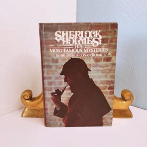 May include: A hardcover book titled "Sherlock Holmes: His Most Famous Mysteries" by Sir Arthur Conan Doyle. The cover features an illustration of Sherlock Holmes smoking a pipe against a brick background. The book is propped up by gold-colored bookends.