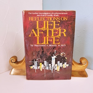 May include: A hardcover book titled "REFLECTIONS ON LIFE AFTER LIFE" by Raymond A. Moody, Jr., M.D. The book has a maroon cover with gold lettering and an abstract illustration. The book is held by gold-colored bookends.