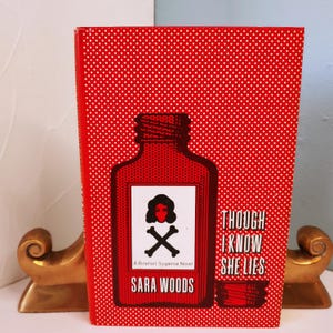 May include: A hardcover book with a red cover and white polka dots. The cover features a red bottle illustration with a skull and crossbones, and the title "THOUGH I KNOW SHE LIES" and author "SARA WOODS".