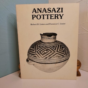 May include: A hardcover book titled Anasazi Pottery by Robert H. Lister and Florence C. Lister. The off-white cover features a large, centered black and white illustration of a round, patterned ceramic vessel with a narrow neck and side handle.