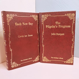 May include: Two hardcover books with burgundy covers and gold lettering. The book on the left is titled "Each New Day" by Corrie ten Boom. The book on the right is titled "The Pilgrim's Progress" by John Bunyan. Both books are from "The Christian Library".