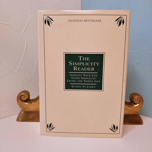 May include: A hardcover book titled "The Simplicity Reader" by Elaine St. James. The book has a cream-colored cover with a dark green rectangle containing the title and subtitle. The words "National Bestseller" are printed at the top.