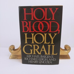 May include: A hardcover book titled Holy Blood, Holy Grail by Michael Baigent, Richard Leigh, and Henry Lincoln. The black cover features the title in large red and gold lettering, supported by two gold-colored metal bookends with scroll details.