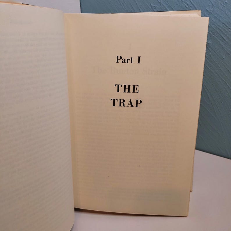 May include: An open book with a cream-colored page featuring the title "THE TRAP" in bold black letters. The text is centered on the page, with "Part I" above the title. The book's pages are slightly yellowed, suggesting age.