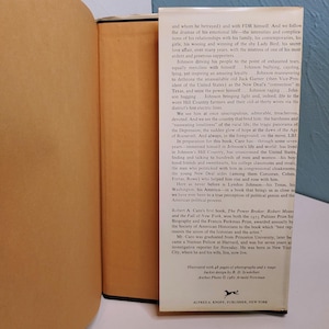 May include: A hardcover book with a cream-colored spine and pages. The title and author's name are not visible. The book's cover features text and is set against a light background. The book is open, revealing printed text.