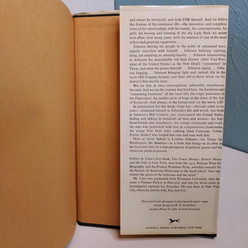 May include: A hardcover book with a cream-colored spine and pages. The title and author's name are not visible. The book's cover features text and is set against a light background. The book is open, revealing printed text.