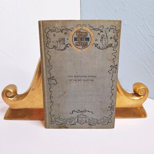 May include: A vintage book titled "Before He Is Twenty: Five Perplexing Phases of the Boy Question" with a gold-colored title and decorative border. The book cover is a light gray color with a textured surface.