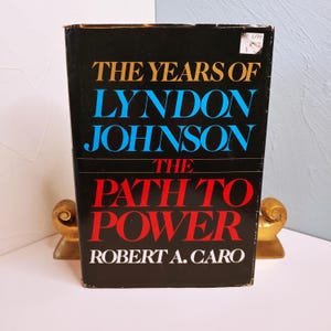 May include: A hardcover book titled "The Years of Lyndon Johnson The Path to Power" by Robert A. Caro. The book has a black cover with gold, blue, and red lettering. The book is propped up by two gold-colored bookends.