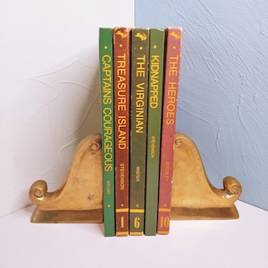 May include: Five hardcover books titled Captains Courageous, Treasure Island, The Virginian, Kidnapped, and The Heroes, held between two gold-toned metal bookends with curved, scroll-like designs on a flat surface.